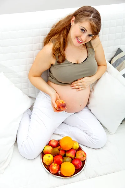 Mujer embarazada sentada en el sofá y sosteniendo la fruta en la mano — Foto de Stock