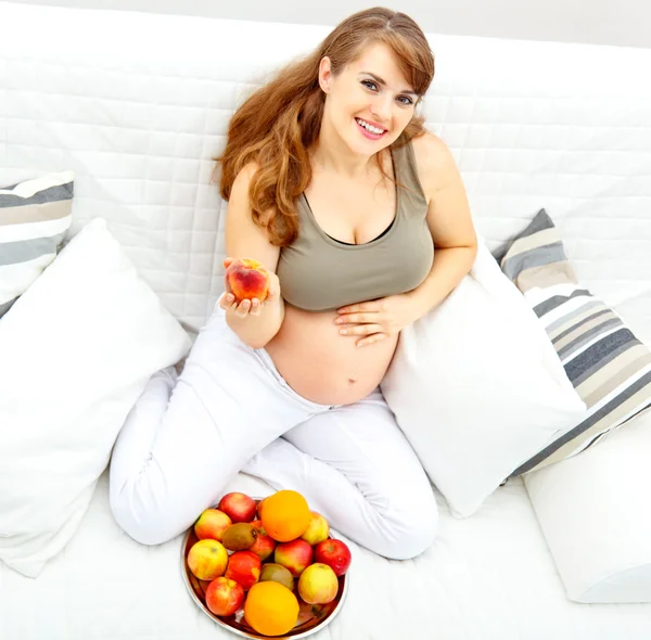 Mulher grávida sentada no sofá e segurando frutas na mão — Fotografia de Stock