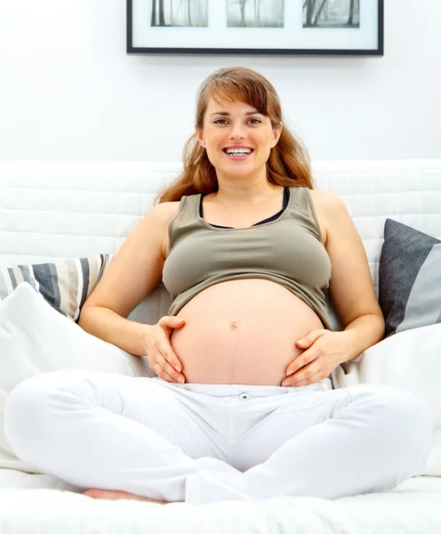 Улыбающаяся красивая беременная женщина сидит на диване и трогает живот. . — стоковое фото