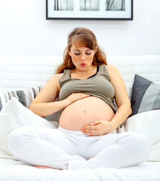 Schöne schwangere Frau sitzt auf dem Sofa und pustet ihren Bauch. — Stockfoto