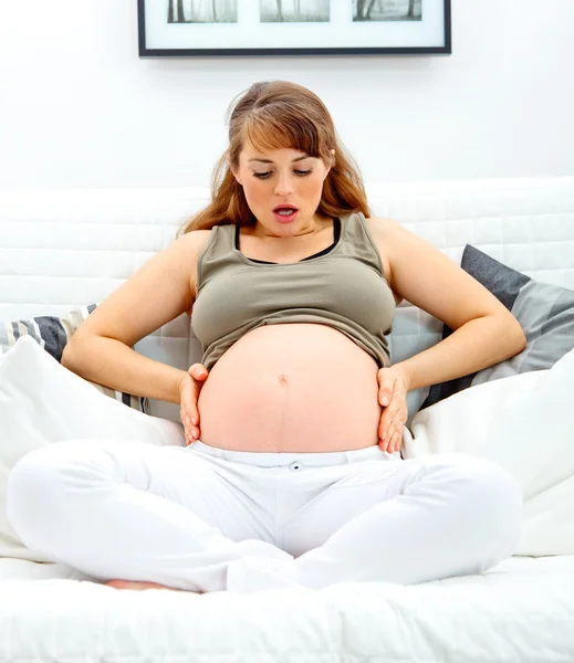 Έκπληκτος όμορφη έγκυος γυναίκα κάθεται στον καναπέ και κρατώντας την κοιλιά. — Φωτογραφία Αρχείου