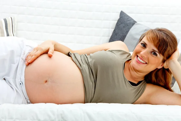 Uśmiechający się piękna kobieta w ciąży na kanapie i przytrzymanie jej brzuch. — Zdjęcie stockowe