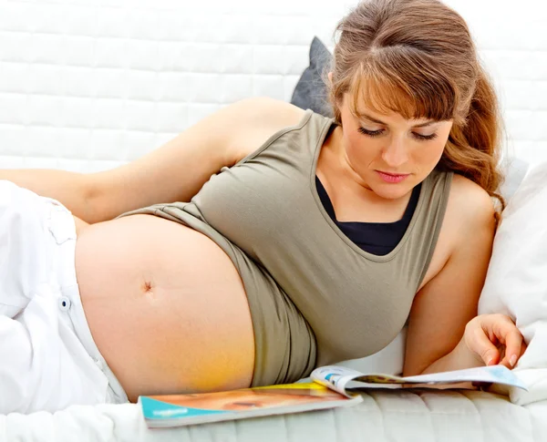 Όμορφη έγκυο γυναίκα ξαπλωμένη στον καναπέ με το περιοδικό — Φωτογραφία Αρχείου