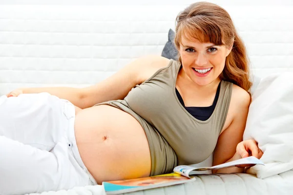 Usmívající se krásná těhotná žena uvolňující na pohovce s časopisem. — Stock fotografie