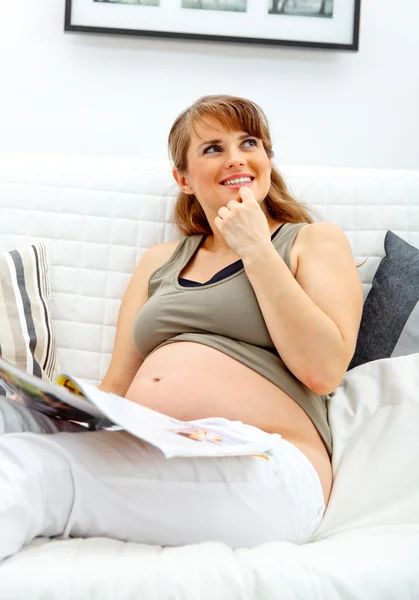 Soñando hermosa mujer embarazada relajarse en el sofá con la revista . —  Fotos de Stock