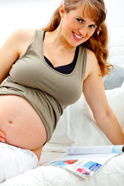 Sorrindo bela mulher grávida sentada no sofá com revista . — Fotografia de Stock