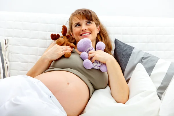 Dreaming beautiful pregnant woman lying on sofa and holding toys. — Stock Photo, Image