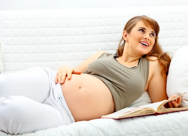 Sorrindo bela mulher grávida relaxando no sofá com livro . — Fotografia de Stock