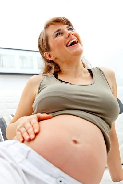 Смеясь красивая беременная женщина расслабляется на диване и держит живот — стоковое фото