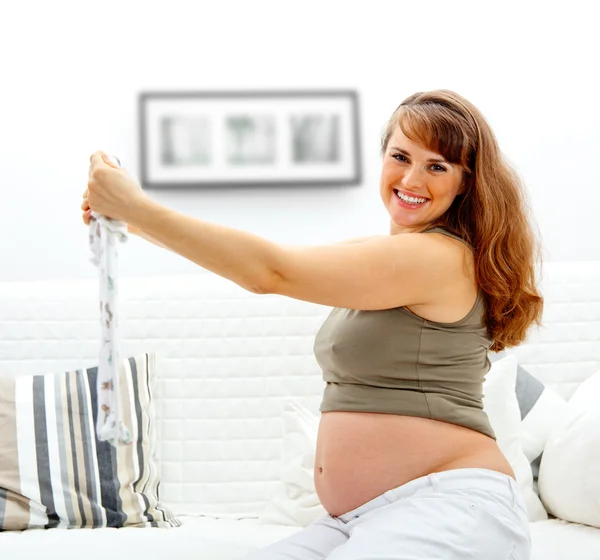 Χαμογελώντας όμορφη έγκυος γυναίκα κάθεται στον καναπέ στο σπίτι με τα ρούχα του μωρού — Φωτογραφία Αρχείου