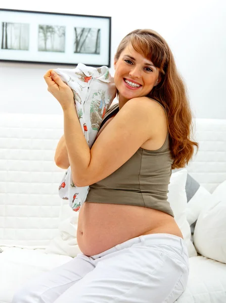 Uśmiechający się piękna w ciąży kobieta siedzi na kanapie w domu z ubrania dla dzieci — Zdjęcie stockowe