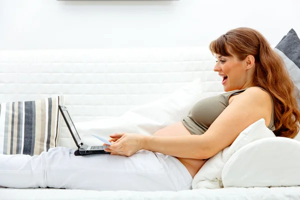 Hermosa mujer embarazada sentada en el sofá con ordenador portátil y tarjeta de crédito —  Fotos de Stock