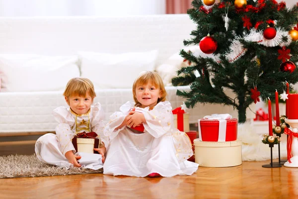 坐在一起的两个快乐的双胞胎女孩提出了圣诞树下 — 图库照片