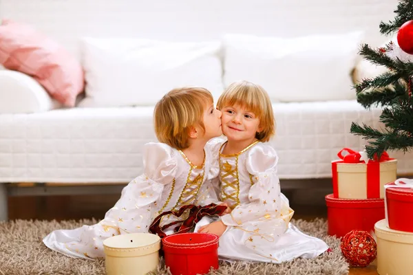 Девушка-близнец целует свою сестру рядом с елкой с подарками — стоковое фото