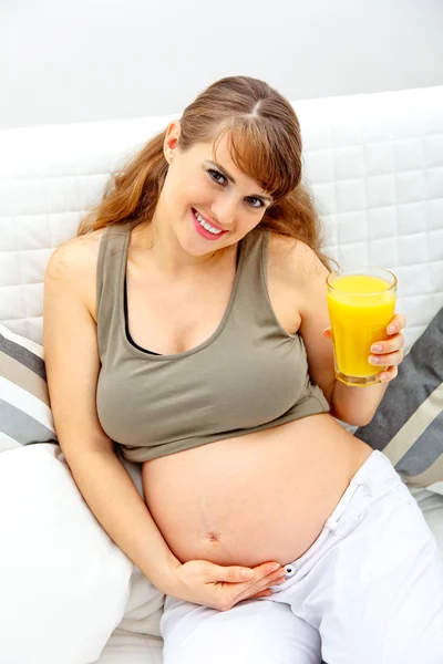 Smiling beautiful pregnant woman sitting on sofa with glass of juice in ha Stock Picture