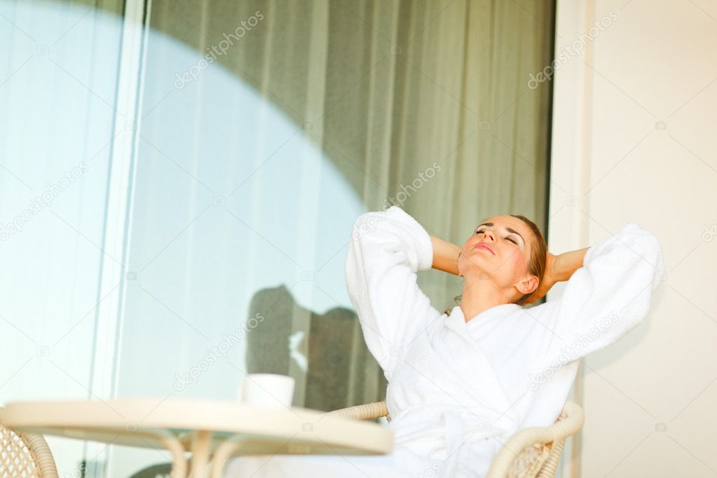 Young female in bathrobe sitting at table on terrace and relaxin