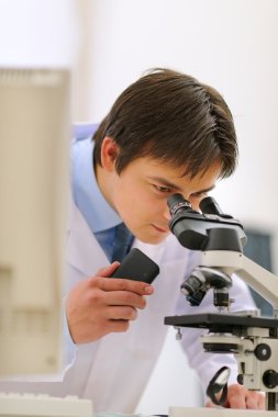 Researcher looking in microscope and making notes on voice recor clipart