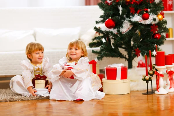 Två glada tvillingar flicka sitter med presentförpackning nära julgran — Stockfoto
