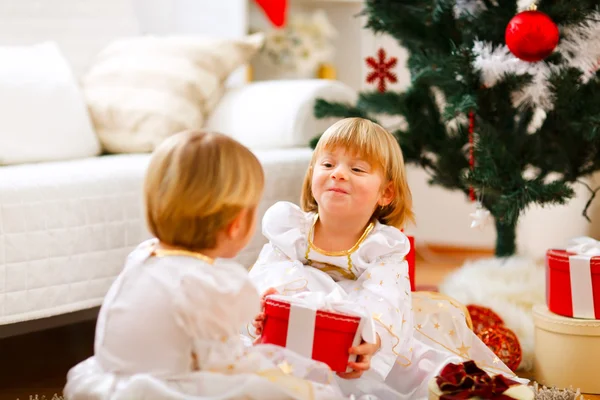 Glückliches Mädchen, das neben dem Weihnachtsbaum sitzt und ihm ein Geschenk macht — Stockfoto