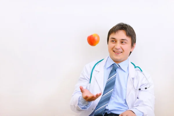 Smiling medical doctor sitting on floor and throwing up apple — Stock Photo, Image