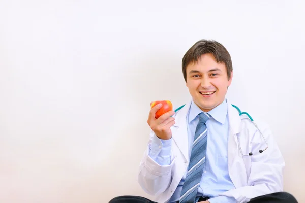 Uśmiechający się lekarz siedzi na podłodze i trzyma jabłko w han — Zdjęcie stockowe