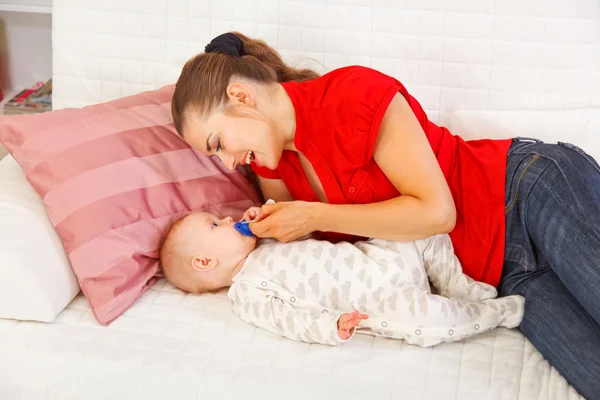 Happy mother laying on couch and giving soother to baby — Stock Photo, Image