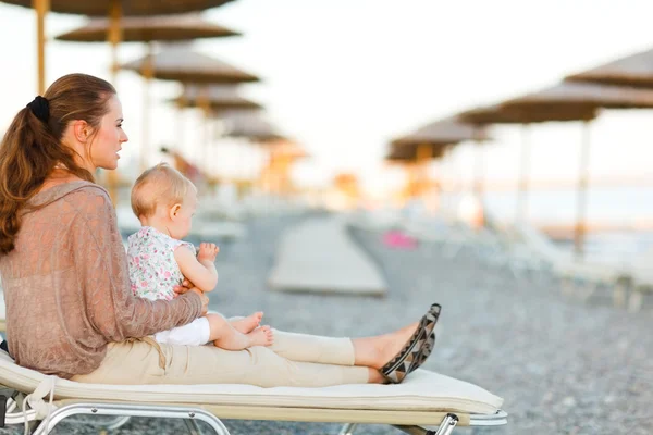 Madre joven sentada con el bebé en la cama de sol en la playa por la noche —  Fotos de Stock