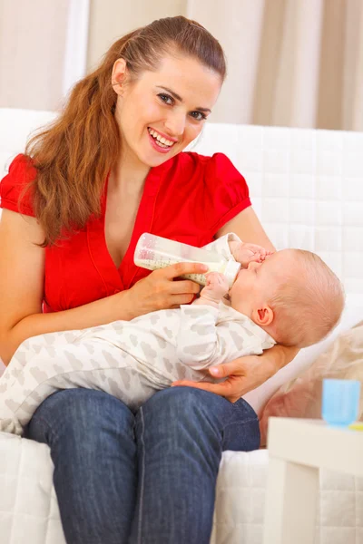 Smiling young mother sitting on couch and feeding baby — Stock Photo, Image