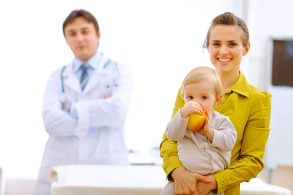 Porträt einer Mutter mit Baby mit Apfel und Doktor im Hintergrund — Stockfoto