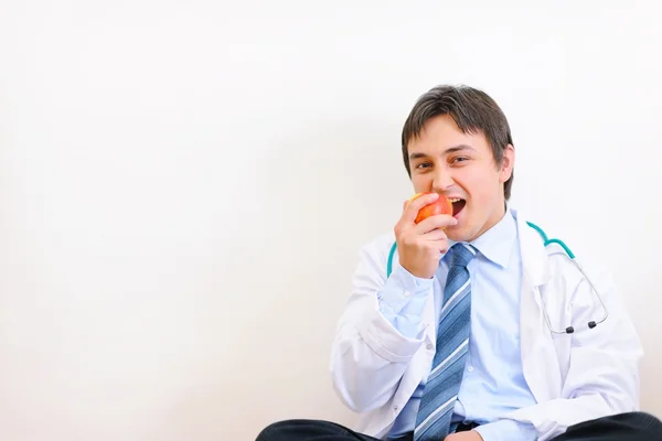 Happy medical doctor sitting on floor and eating apple — Stock Photo, Image