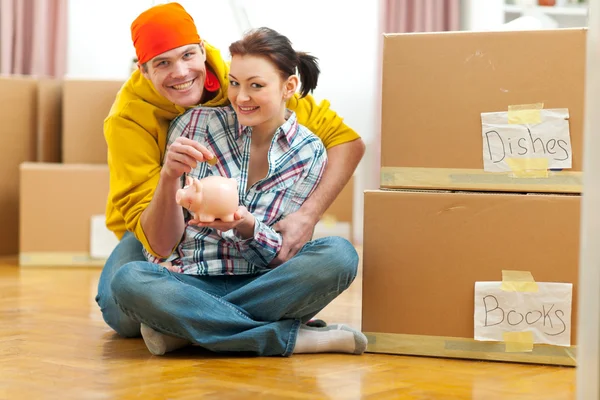 Young couple among boxes putting coin in piggy bank — Stock Photo, Image