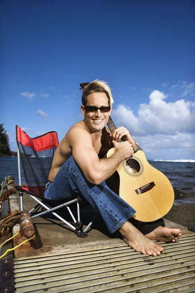 Fiatal ember ül tóparti és a gazdaság gitár — Stock Fotó