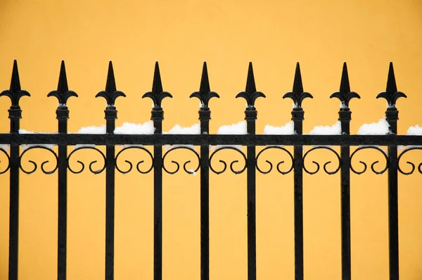 Old fashioned spike fence against yellow wall, some snow — Stock Photo, Image