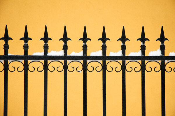 Old fashioned spike fence against yellow wall, some snow