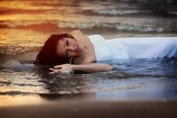 Jovem atraente no vestido de noiva goza de água do mar durante o pôr do sol Imagem De Stock