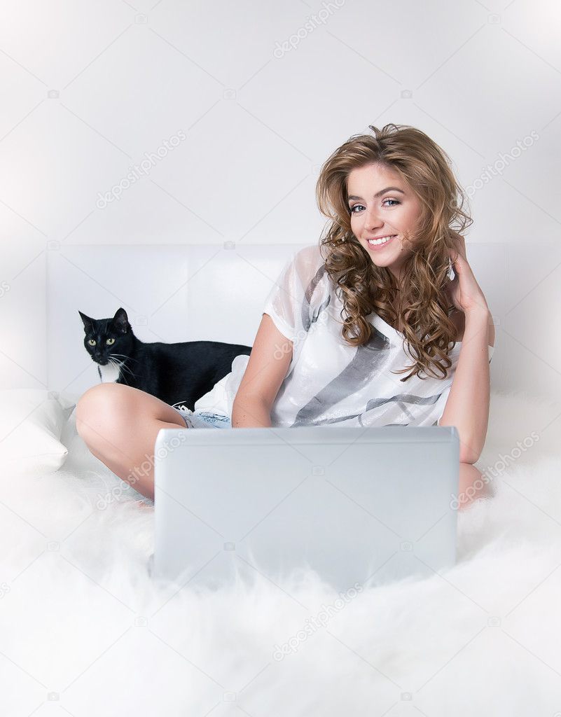 Smiling young girl in white bed with black cat and laptop