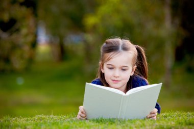 A little girl enjoying reading outdoors on the grass clipart