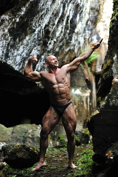 Young bodybuilder posing in a cave - front view in full length p — Stock Photo, Image