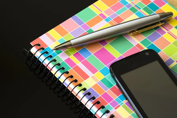 Telefone celular preto com notebook colorido e caneta de prata isolado no fundo preto — Fotografia de Stock