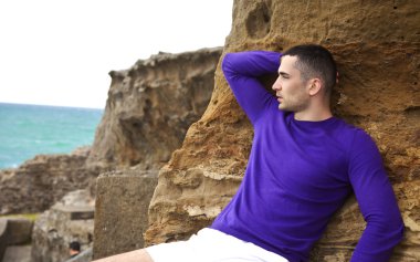 Man laying on the rocks on the beach looking toward the sunlit water pensiv clipart