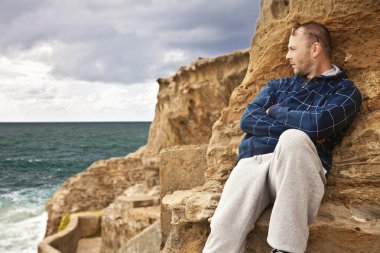 Man laying on the rocks on the beach looking toward the sunlit water pensiv clipart