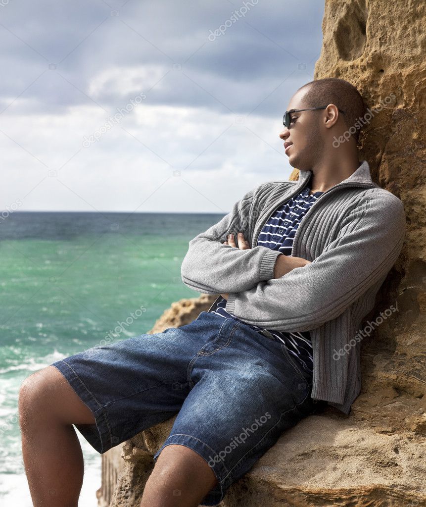 Black man laying on the rocks on the beach looking toward the sunlit water