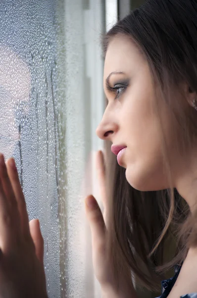 Belle fille regardant par une fenêtre humide Image En Vente