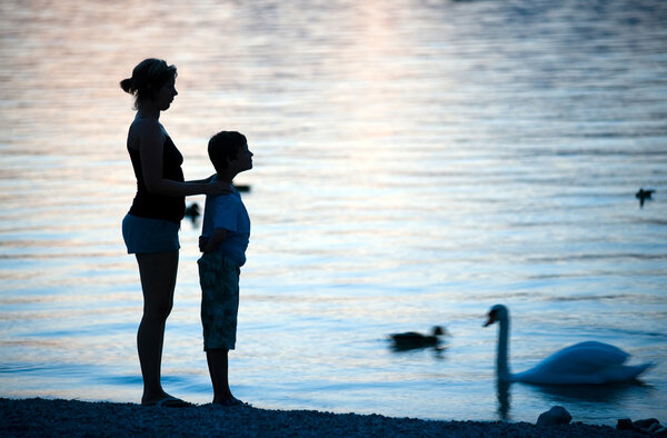 A mother and her son are watching swimming ducks and swans on a lake during dawn.