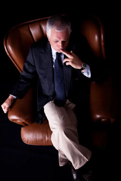stock image Pensive mature businessman seated on a chair, isolated on black background. Studio shot.