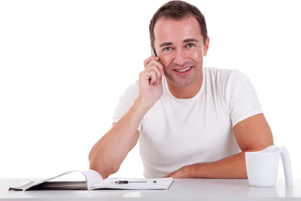 Sorrindo homem de meia-idade sentado na mesa no telefone, em um fundo branco. Estúdio — Fotografia de Stock