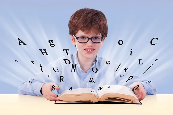 Cute boy reading a book on his desk, with flying letters, isolated on blue, studio shot — Stock Photo, Image