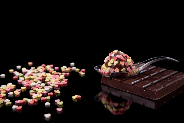 Schokolade - Brigadier auf einem Löffel, mit Nuggets herzförmig, farbig, auf schwarz mit Reflexion Stockbild