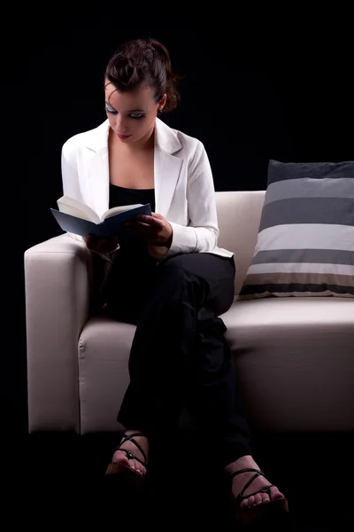 Beautiful woman seated on couch reading a book, isolated on black. Studio shot. — Stock Photo, Image