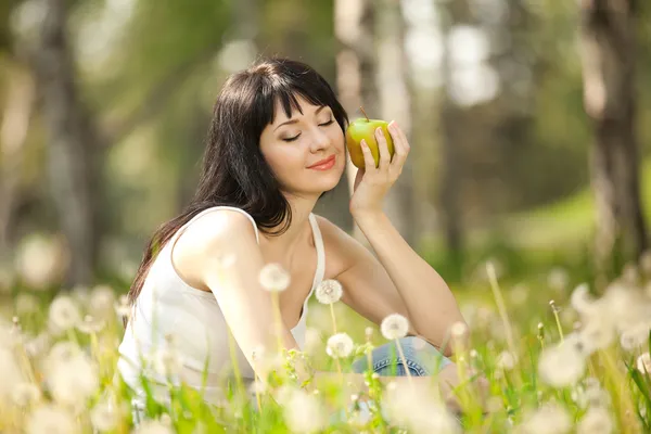 Mujer linda comiendo la manzana en el parque con dientes de león —  Fotos de Stock